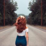 Red-haired woman in jean shorts walking down a forest road, showcasing style and freedom.